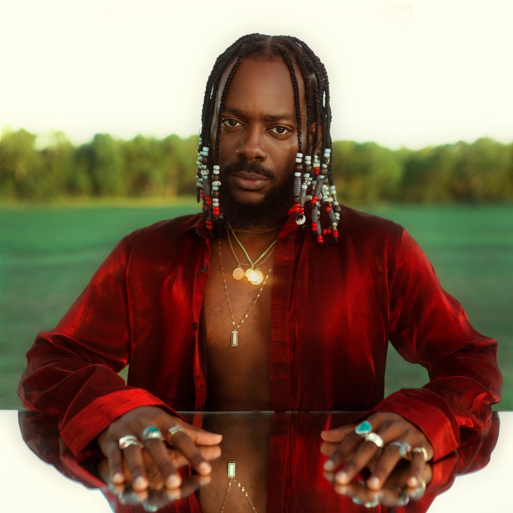 Adekunle Gold in a red satin shirt, seated at a mirrored table