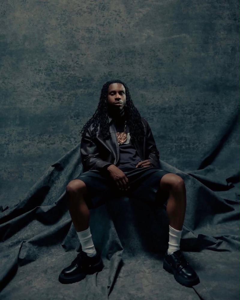 Polo G seated in a leather jacket against a dark, textured backdrop
